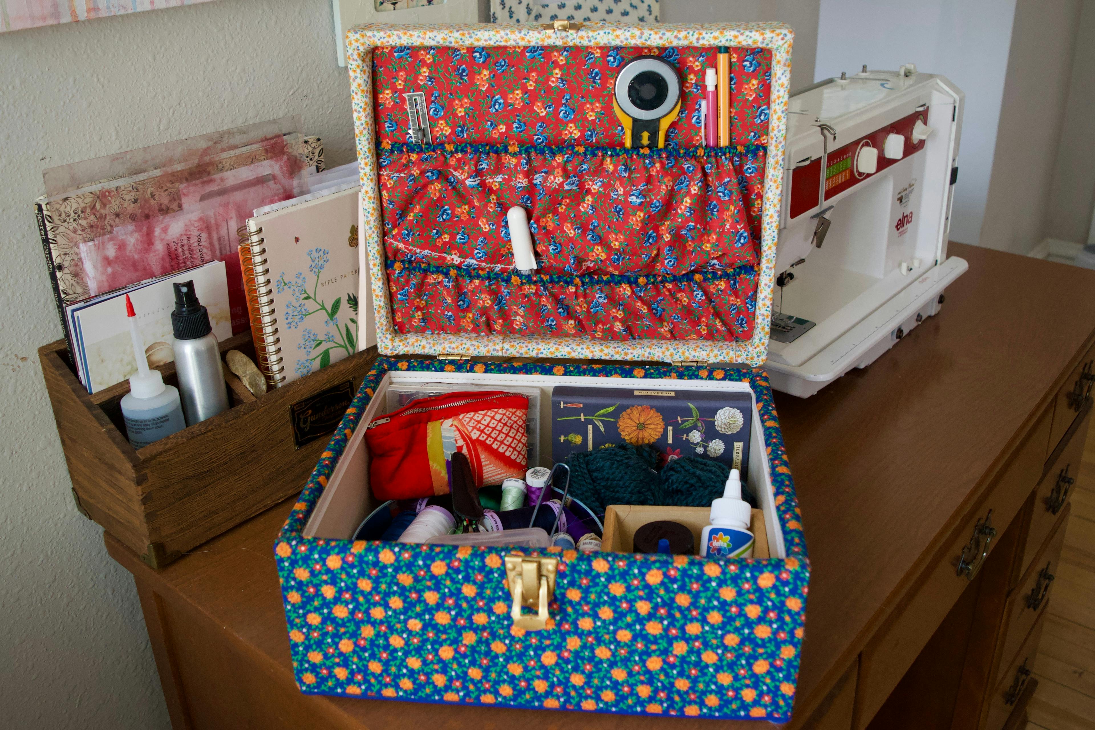 A sewing box sitting open showing its contents. The box is on a sewing table with a sewing machine and wooden containers of stationary and other crafting supplies.