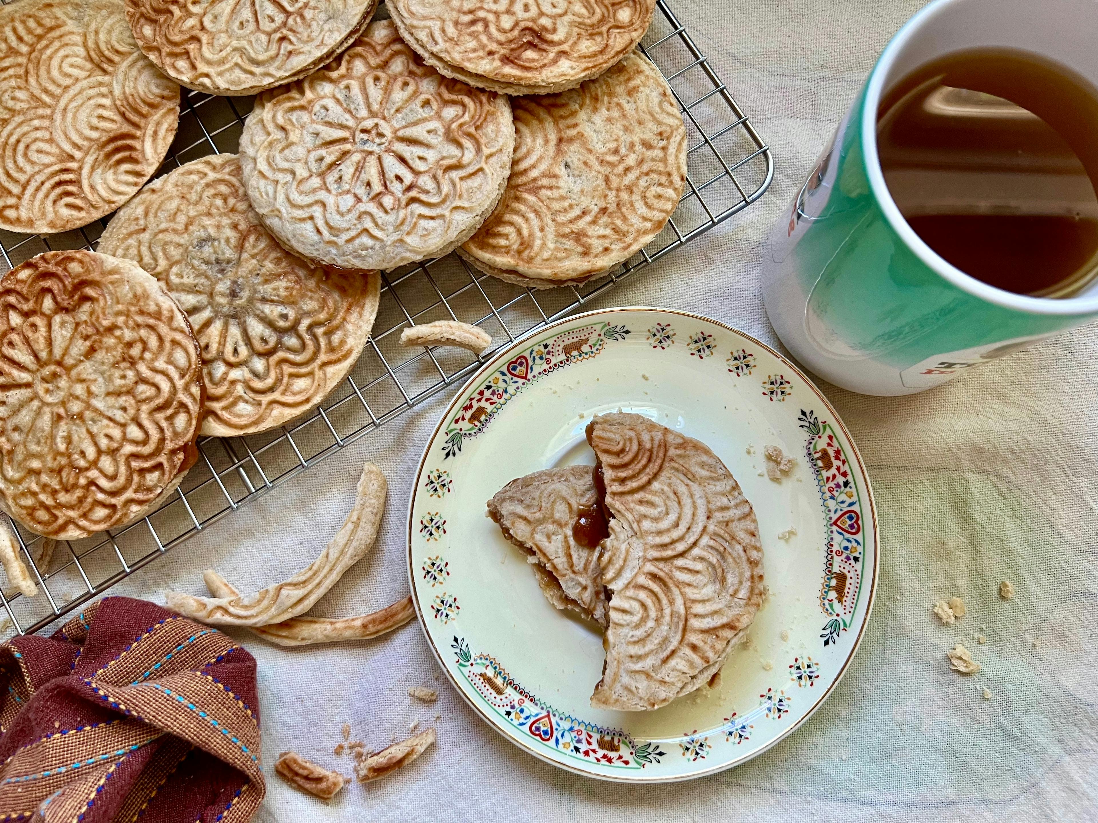 A bird's eye view of a wire rack of stroopwafels, plus one stroopwafel broken in half with caramel oozing out on a small saucer. There is also a cup of tea in frame.