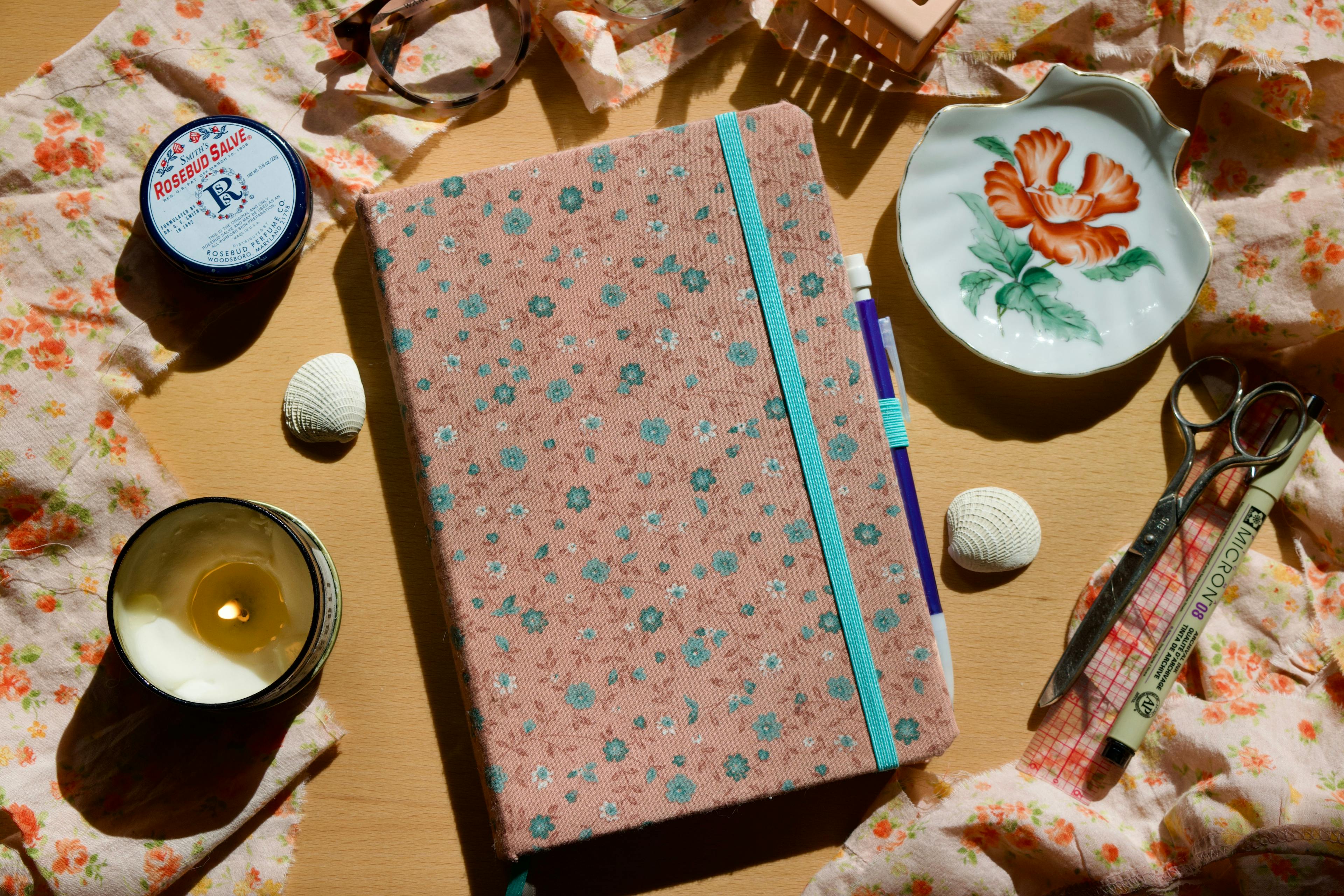 A journal is lying on a wooden table. Its cover is a pink vintage fabric with tiny blue flowers. The journal is held closed by a minty green elastic band and there are two ribbon bookmarks, one the same minty green and one grey. It is surrounded by various desk items such as a candle, glasses, pens, lip balm, a small porcelain dish, and a scrunch of different pink floral fabric. 