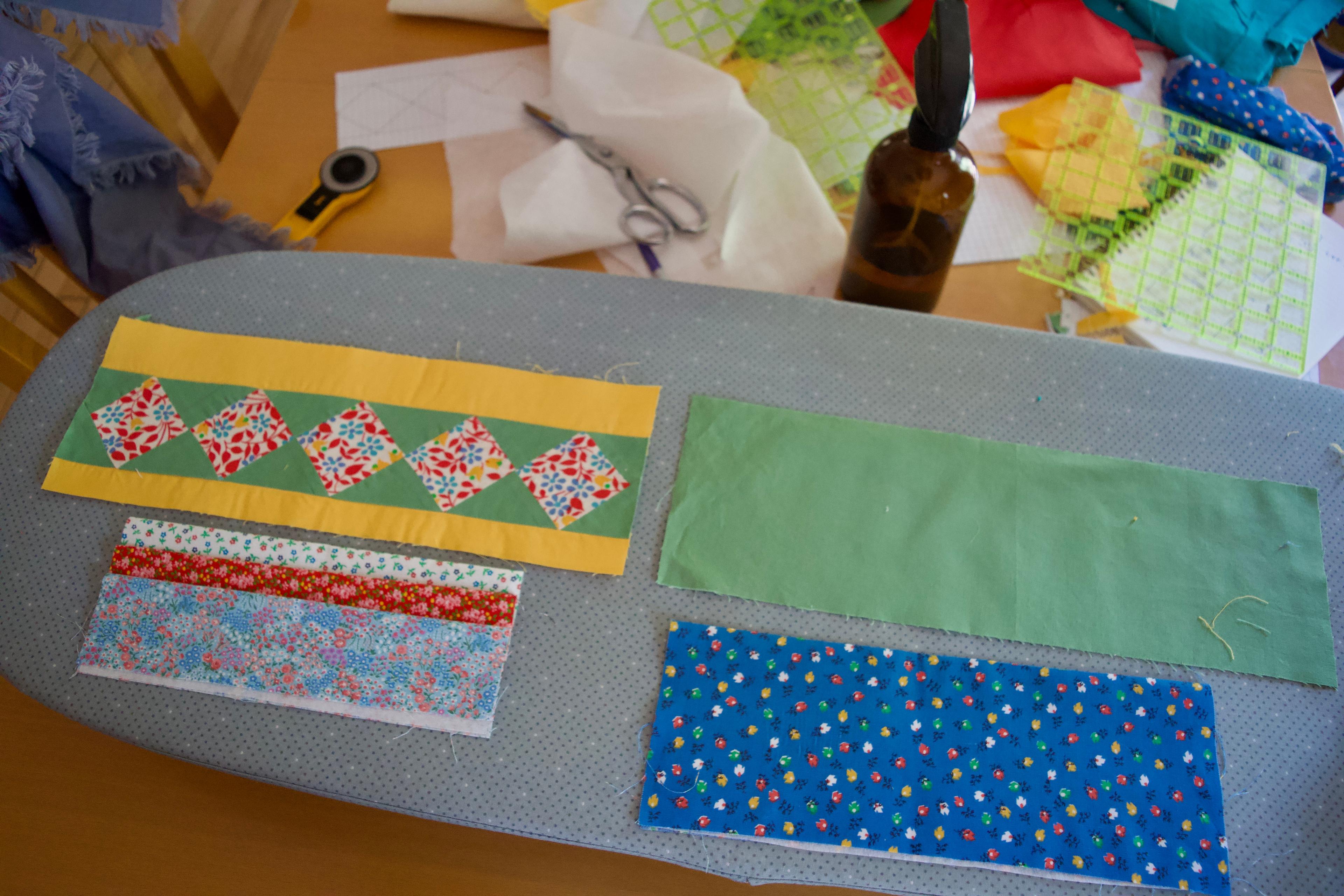 All of my pieces laid out on an ironing board - the outside patchwork piece included. There is a mess of fabrics, cutting utensils, and rulers on the table in the background.