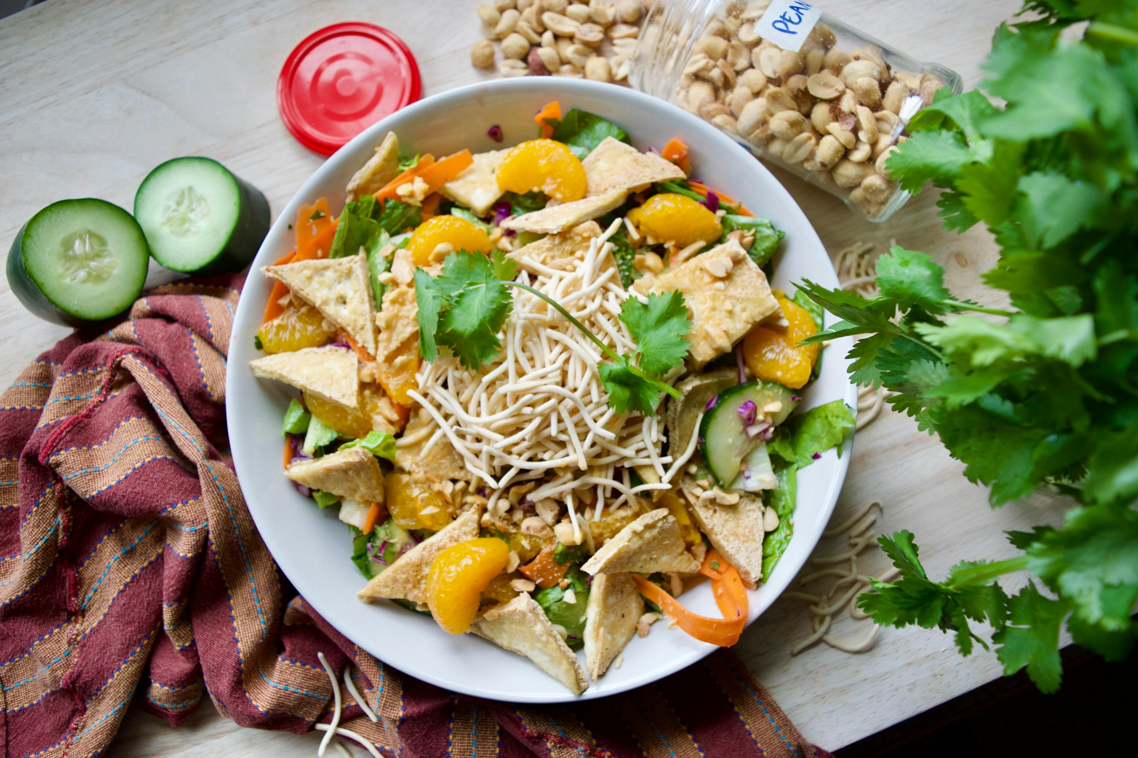 A large plate of a salad artfully surrounded by some of the ingredients within such as cucumbers, cilantro, and peanuts. The salad visibly contains carrots, chow mein noodles, fried tofu, and mandarin oranges.
