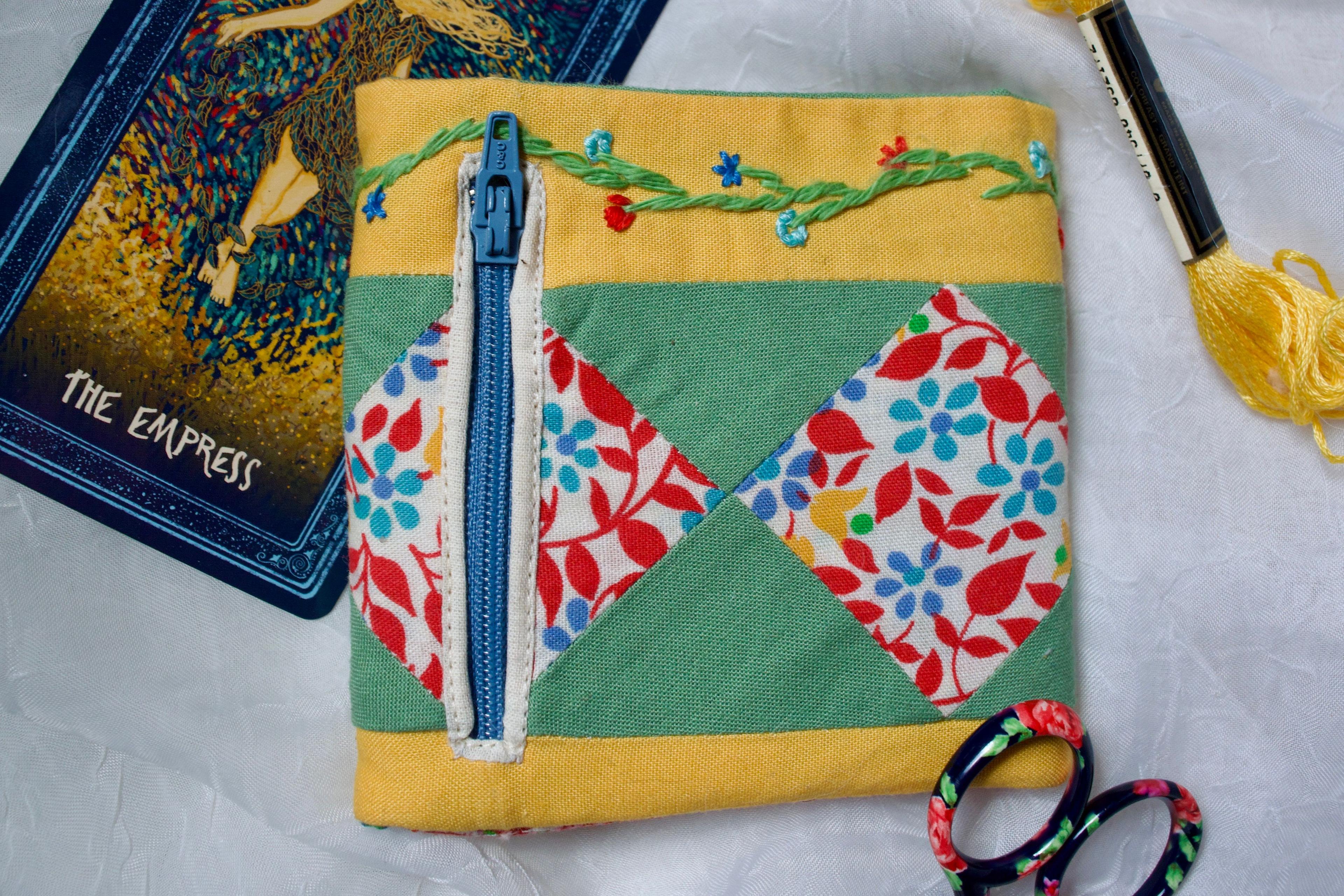 A fabric wallet with floral embroidery sits on a white piece of fabric. The wallet is mostly yellow and green, with diamonds of a floral fabric that has red, yellow, and blue flowers. The zipper for a small coin pocket is visible and also blue. There is The Empress tarot card, yellow embroidery floss, and a small pair of scissors also visible. 